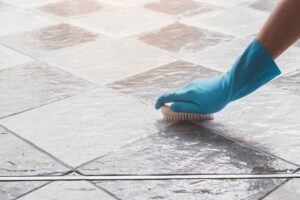 Mano con guanto azzurro e spazzola su pavimento per pulire fughe